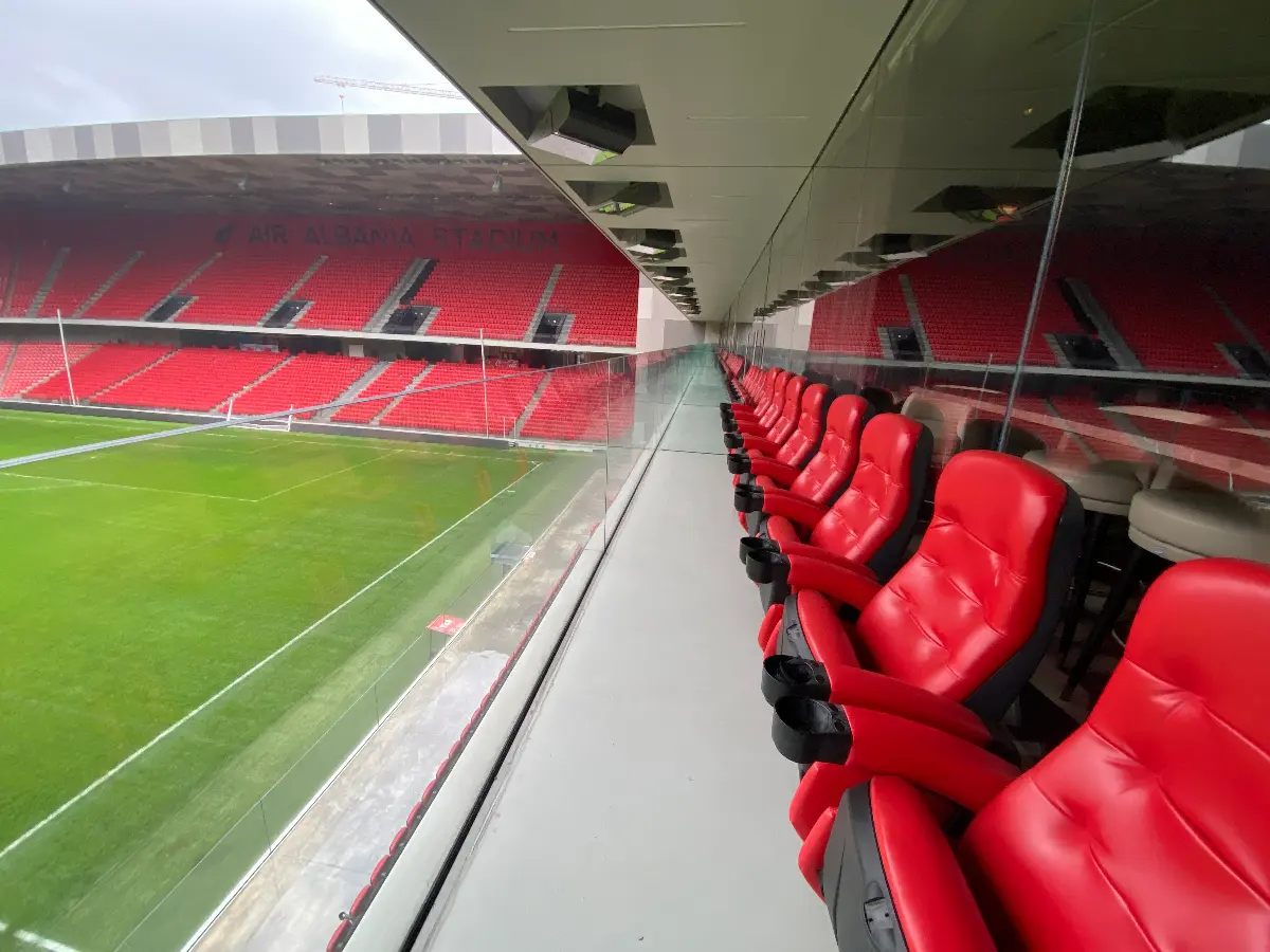 Skybox met comfortabele rode stoelen in het Air Albania Stadion, met uitzicht op het groene speelveld en de rode tribunes van het stadion.