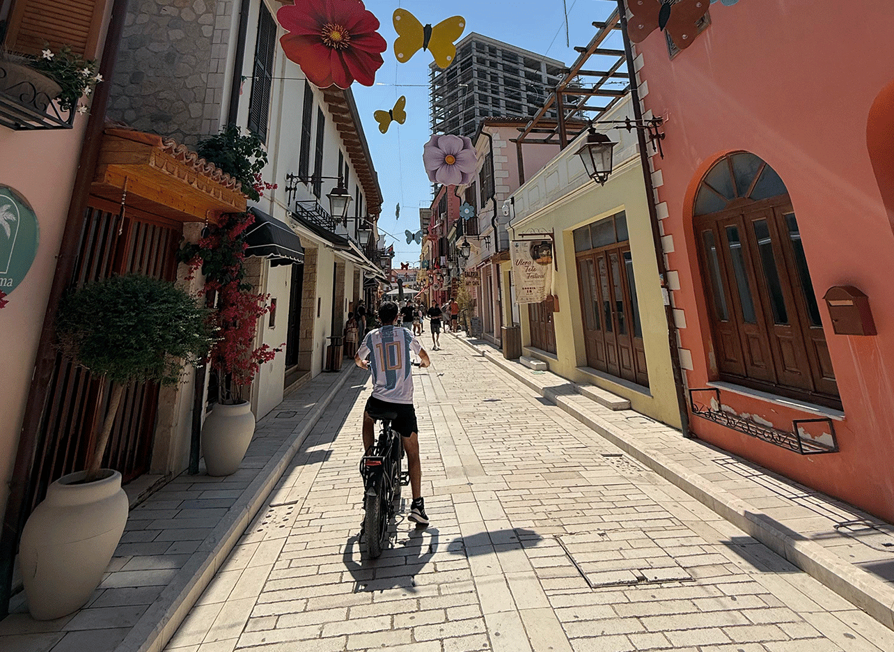 Sfeervolle straat met winkels en hangende bloemen en vlinders, mensen wandelen en fietsen.